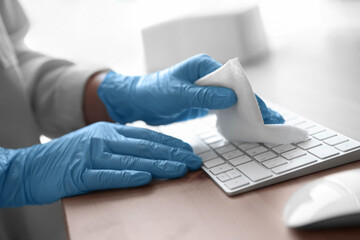 Woman in latex gloves cleaning computer keyboard with wet wipe at table, closeup