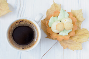 Cup of freshly brewed black coffee on a wooden white background next to cupcake on autumn maple leave as a tray, top view
