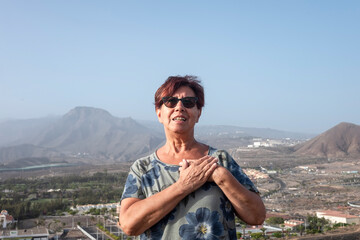 Naklejka premium Mature woman standing on hill with landscape of mountains and city on background looking at camera with hands on the heart - active senior and freedom concept