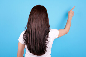 Brunette girl with long hair pointing with finger, view from behind. Isolated on blue background.