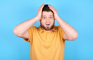 Brunette man holding his hands on head, shocked emotion. Isolated on blue background.