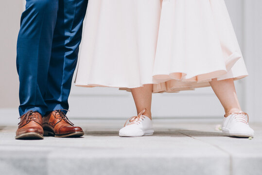 Bride and groom stand against each other. Groom with leather brown lacquered shoes, blue suit pants and the bride with white pink dress. Concrete pavement. Outdoor wedding, celebration and good mood.