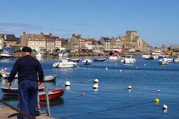A fisherman walking beside a harbour with the village houses of Barfleur, France, in the background.