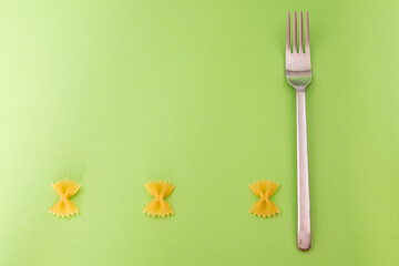 Short raw butterfly-shaped pasta draws a horizontal line leaving ample room for text on the green background, a steel fork is placed on the side.
