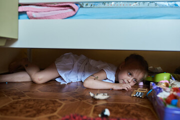 A girl hiding under a bed with toys. Girl in white dress lies on the floor under the crib