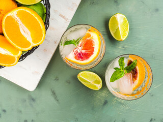 Fresh summer citrus lemonade with lime, mint, blood oranges and ice cubes in jar and glasses over green textured background. Flat lay