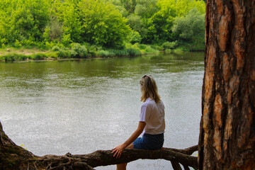 Rear View of a dreaming girl sitting on a tree in the forest by the river in the background.