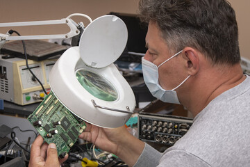 A 40-45-year-old man in a medical mask looks through a magnifying glass at an electrical Board from a computer. Repair of office equipment at home.