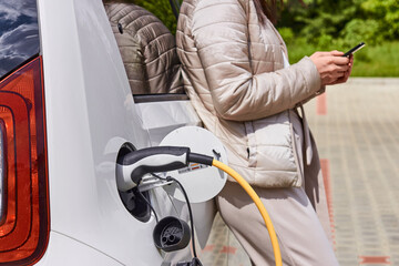 Woman with a mobile phone near recharging electric car. Vehicle charging at public charging station...