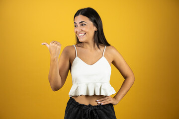 Young beautiful woman over isolated yellow background pointing thumb up to the side smiling happy