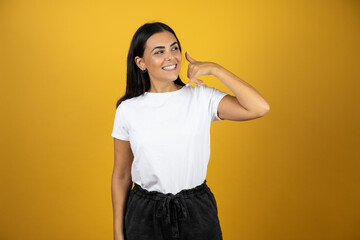 Young beautiful woman smiling and putting her hand next to her ear like it was a telephone. Wearing casual white t-shirt on yellow background