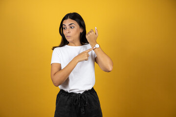 Young beautiful woman standing over isolated yellow background surprised and pointing his watch...