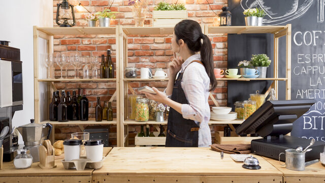side view of young girl waitress in apron looking back on shelf finding tools of making coffee in counter. female barista searching for cup and thinking while holding note of customer order in cafe