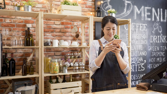 beautiful young female barista in apron is using smartphone and smiling while standing in bar counter at cafe store. elegant lady staff of coffeehouse relax in workplace chatting online on cellphone.