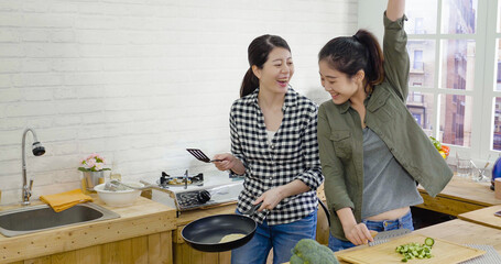 two asian korean best friends making homemade food in modern kitchen. cheerful ladies laughing and having fun while preparing snack for tea time. young girl successful flipping pancake on frying pan