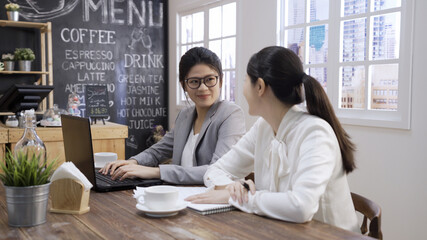 asian japanese female graphic designer typing on laptop computer during collaboration with women colleague on common project in coffee shop. Two lady coworkers discussing website on notebook pc.