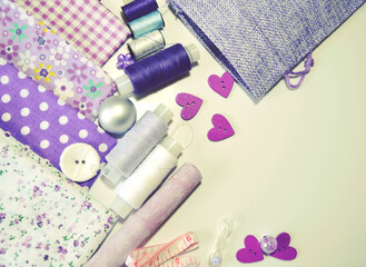 Flat lay of sewing accessories including white-dotted and floral cotton fabrics, buttons, piece of chalk,  sewing spools in lilac and mauve against ecru background. Scrapbooking and DIY. Hobby