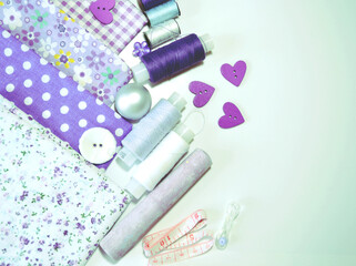 Flat lay of sewing accessories including white-dotted and floral cotton fabrics, buttons, piece of chalk, sewing spools in lilac and mauve against white background. Scrapbooking and DIY. Hobby