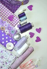 Fototapeta na wymiar Flat lay of sewing accessories including white-dotted and floral cotton fabrics, buttons, piece of chalk, sewing spools in lilac and mauve against white background. Scrapbooking and DIY. Hobby