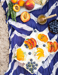 Obraz na płótnie Canvas Healthy picnic for a summer vacation on the sea beach with fresh fruits and berries, mini sandwiches with cream cheese, fruits and blueberries with honey on a blue and white cloth.