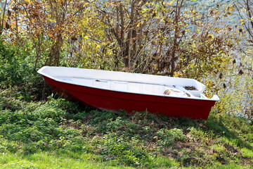 Very small plastic white and red river boat left on side of river bank surrounded with grass and dense branches on warm sunny autumn day