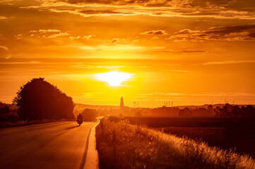 Naklejka na ściany i meble Beautiful scene of a main road in the sunset ending in the horizont with Nördlingen in Bavaria Germany. A motorbike is driving down the road towards the suns.