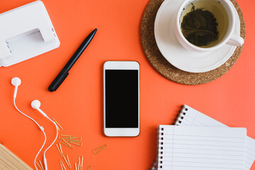 Mobile phone, cup of green tea, notebooks, pen, paper clips, hole punch and earphones on orange background, flat lay. Back to school