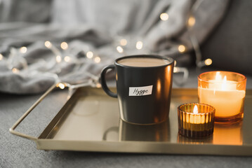 Hygge concept: hot coffee in black mug served on a metal tray with two burning candles, decorative lights as background. Cozy atmosphere