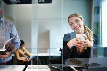 Portrait of smiling young blond businesswoman holding cup while standing by colleague in office seen through glass shield
