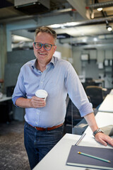 Portrait of smiling mature businessman holding disposable cup while standing by desk at creative office