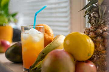 Fruits lie on a windowsill lit by the sun: pineapple, bananas, lemon, orange, apple, mango. In a glass of fruit juice with ice