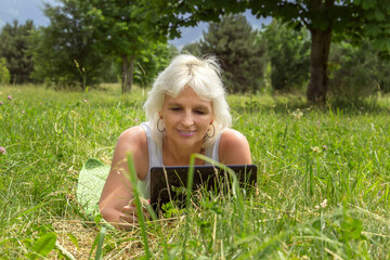 Naklejka na ściany i meble Slender elderly woman with gray hair lies in the park on the grass and communicates via laptop
