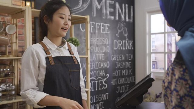 islam woman customer paying credit card at cafe store. young asian smiling barista in apron giving client order of coffee in paper cup and using point of sale terminal with payment from arabic lady