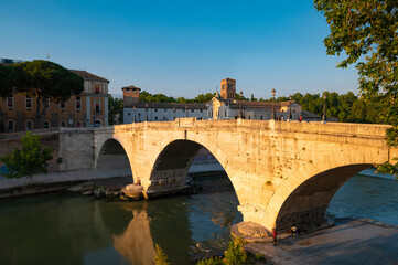 Obraz premium Ponte Fabricio, Rome, connects the Tiber island on the Tiber to the Jewish quarter. View of the arches reflecting in the tiber near the dam on a summer day with blue sky.