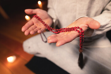 close-up man in clothes for practice and meditation sits in a lotus pose and holds red rosary to concentrate attention in a wooden room with dim light