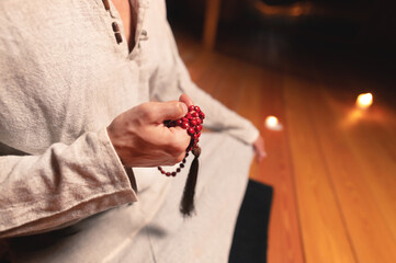 close-up man in clothes for practice and meditation sits in a lotus pose and holds red rosary to...
