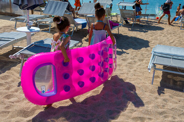 Bambine tra ombrelloni e lettini che si portano il materassino in spiaggia.