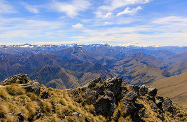 View of the Southern Alps from the climb up Ben Lomond. Tussocks and rocks in the foreground and snow-capped mountains in the background.