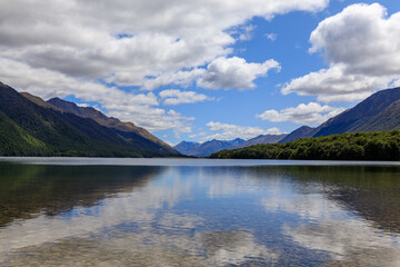 The calm waters of South Mavora Lake with forested mountains down each side and mountains in the distance. White Fluffy clouds on a blue sky.