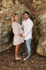 Pregnant couple touching their bellie in a forest and looking at the camera. Blonde pregnant woman with her brunette boyfriend. Blonde pregnant woman in a beige dress.