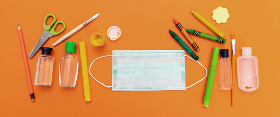Coronavirus COVID 19 spread prevention measure at school. Surgical mask, alcohol disinfection gels and stationery on orange color background,