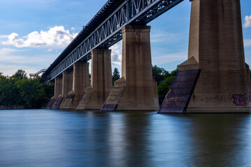 a view of the train bridge in the City of Saskatoon from below on the shore of the South Saskatchewan River. a long exposure time was used to smoothen the surface of the water in the river