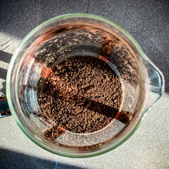 Rich, Brown Coffee grounds in a French Press as seen from above with rays of morning sunlight reflecting through glass.