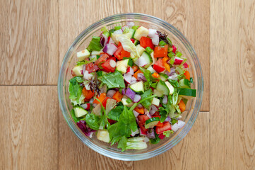 Fresh and healthy vegetables for salad in a glass bowl placed on brown wooden surface.