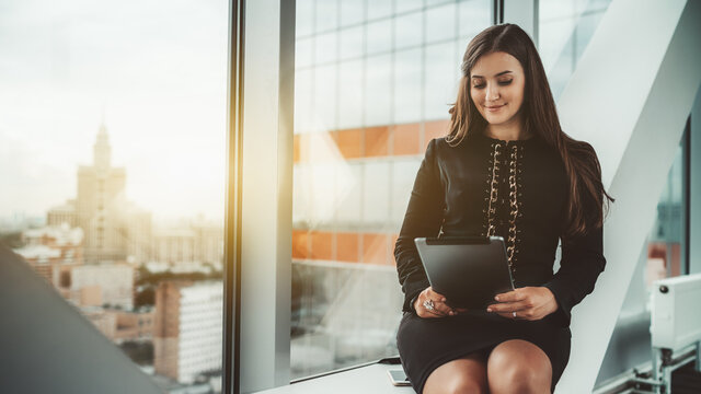 A dainty hispanic businesswoman is sitting on a modern white bench of a business high-rise coworking office area near a panoramic window and using her digital tablet; a copy space place on the left