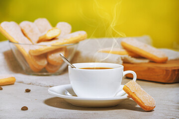 White cup of hot aromatic coffee with steam, traditional italian savoiardi biscuits or ladyfingers cookies on a plate and in a glass bowl. Selective focus