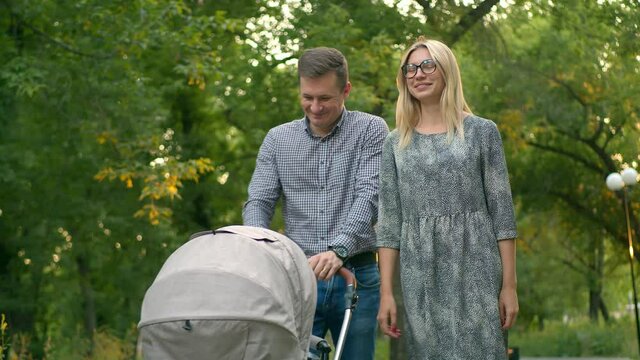 Happy hetero couple walks with a newborn baby in a stroller in the Park. A man and a girl walk along the alley of the Park with a baby stroller having fun and laughing