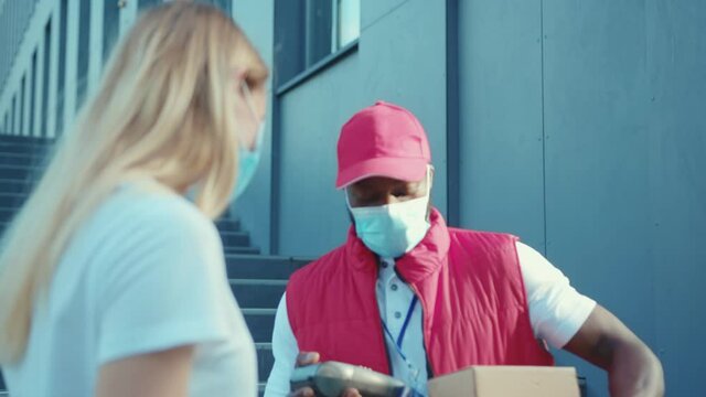 African american man courier wearing a mask delivers a parcel to woman client paying by contactless terminal for parcel transporting payment uniform express order worker outdoors close up slow motion