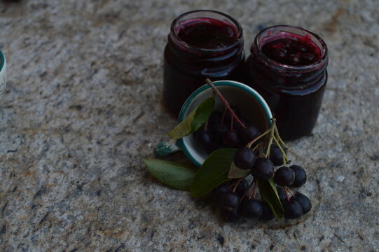 Aronia jam next to fresh berries. Homemade Aronia jam in a glass jar with fresh Aronia berries in cups.
