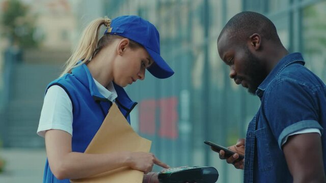 Smiling young woman courier in blue hat hold package african american client paying by phone for parcel transporting payment uniform express order worker outdoors close up slow motion